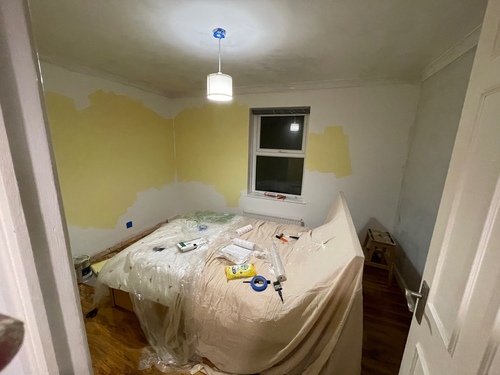 Looking into the partially painted bedroom through the door -- you can see three walls, with the window on the far side. The bed is in the middle of the room, covered in dust sheets and a variety of painting tools. Two of the walls have some patches of the light yellow on them, some of my initial roller work. Outside the window is darkness -- I was painting at night.